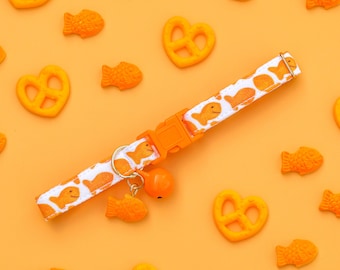 Puede incluir: Un collar blanco para gatos con un estampado de peces naranja y una hebilla naranja. El collar tiene una pequeña campana naranja y un dije en forma de pez. El fondo es naranja con peces y pretzels naranjas dispersos.