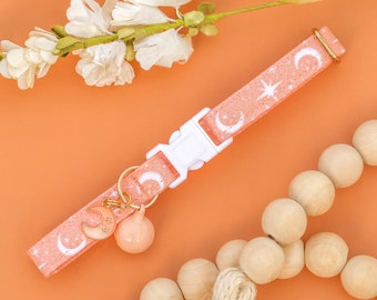 May include: A peach-colored cat collar with a white crescent moon and star pattern. The collar has a white plastic buckle and a gold bell charm with a crescent moon and star design.