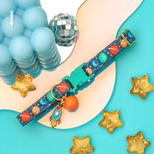 May include: A turquoise blue dog collar with a colorful space-themed pattern. The collar has a rocket ship charm and a bell. The collar is on a blue background with gold stars.