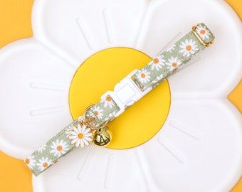 May include: A green and white daisy print cat collar with a gold bell charm. The collar is on a white daisy-shaped background.