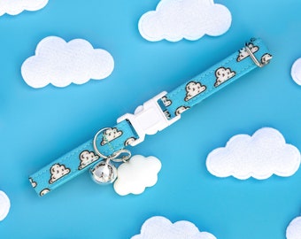 Puede incluir: Un collar de gato azul con una hebilla blanca y una campanilla plateada. El collar tiene un patrón de nubes blancas sobre un fondo azul. El collar está sobre una superficie azul con decoraciones en forma de nube blanca.