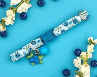 May include: A blue and white patterned cat collar with a bell charm. The collar has a floral design with blue flowers and white leaves. The bell is gold and has a blue bell.