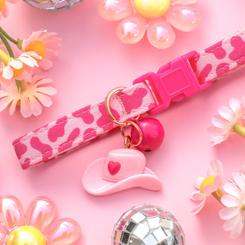 May include: Pink and white patterned pet collar with a pink heart-shaped cowboy hat charm and a pink bell. The collar is on a pink background with pink flowers.