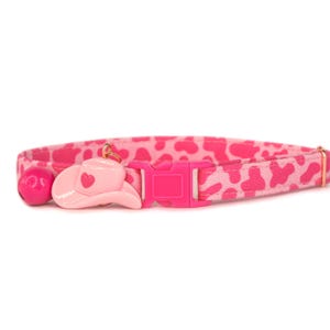 May include: Pink cat collar with a pink cowboy hat charm and a bell. The collar has a pink and white leopard print pattern.