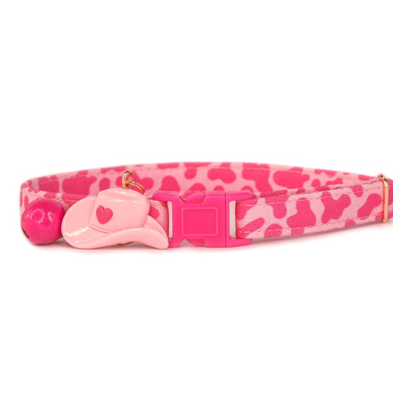 May include: Pink cat collar with a pink cowboy hat charm and a bell. The collar has a pink and white leopard print pattern.