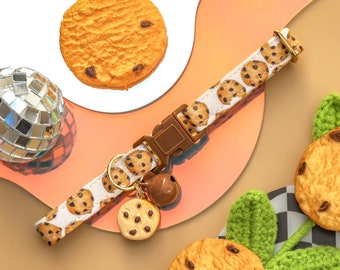 Può includere: Un collare per gatti bianco con una fibbia regolabile marrone e un campanello. Il collare è decorato con un motivo di biscotti con gocce di cioccolato.