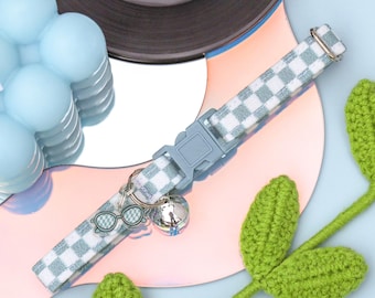 Puede incluir: Un collar de gato a cuadros azul claro y blanco con una campanita plateada y un dije con forma de gafas de sol.