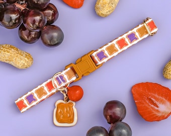 May include: A colorful dog collar with a peanut butter and jelly sandwich charm. The collar is decorated with purple, orange, and white stripes. The charm is a brown and yellow peanut butter and jelly sandwich.