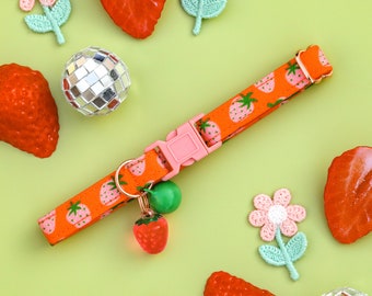 Puede incluir: Un collar de perro con estampado de fresas rojas, blancas y verdes con una hebilla rosa y dos dijes. Un dije es una bola verde y el otro es una fresa roja. El collar está sobre un fondo verde con fresas rojas, flores rosas y bolas de discoteca plateadas.