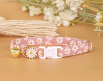May include: A pink cat collar with a white daisy print and a gold bell. The collar has a white plastic buckle.