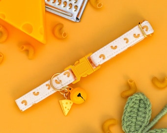 Puede incluir: Un collar de gato blanco con un patrón de macarrones amarillo y un amuleto de campana amarillo. El collar está sobre un fondo amarillo con otras piezas de macarrones y una planta de crochet verde.