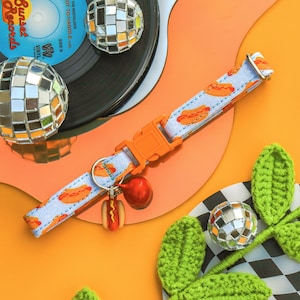 May include: A colorful dog collar with a hot dog print and a small hot dog charm. The collar is orange and white with a red and white hot dog print. The charm is a small, yellow hot dog with mustard and ketchup.