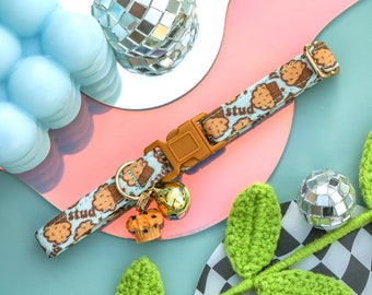 May include: A blue and brown patterned cat collar with a gold bell charm and the word "stud" printed on the collar. The collar is lying on a pink and blue surface with a disco ball and green crocheted leaves.