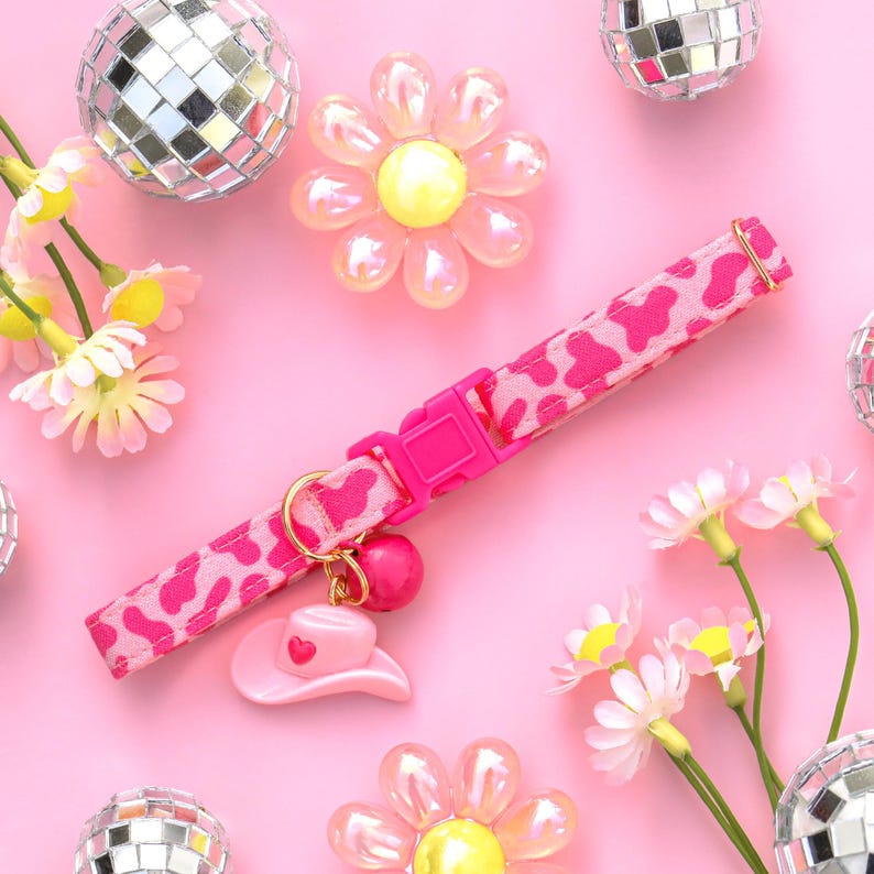 May include: Pink cat collar with a pink cow print pattern and a pink cowboy hat charm. The collar has a pink plastic buckle. The collar is surrounded by pink flowers and silver disco balls.