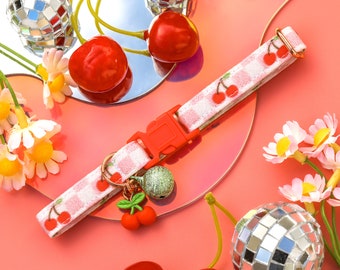 May include: A white and red gingham dog collar with a cherry charm and a bell. The collar is on a pink surface with cherries and a silver disco ball.