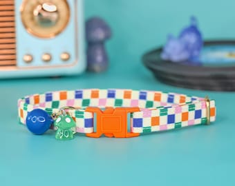 Pode incluir: Um colar de gato xadrez colorido com uma fivela laranja e dois pingentes: um sino azul e um cogumelo verde.