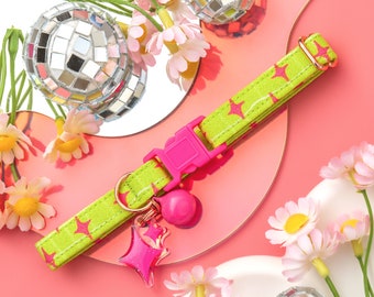 Puede incluir: Un collar de gato verde lima con estrellas rosas y una campana rosa. El collar tiene una hebilla rosa y un amuleto de estrella rosa. El collar está sobre un fondo rosa y blanco con bolas de discoteca y flores.