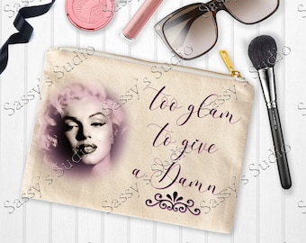 May include: A beige canvas makeup bag with a zip closure. The bag features a black and white image of Marilyn Monroe with the text "too glam to give a Damn" in black cursive lettering.