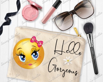 May include: A beige canvas makeup bag featuring a smiling yellow emoji with pink eyelashes and a pink bow. The bag has the text "Hello Gorgeous" in black cursive with a white daisy.