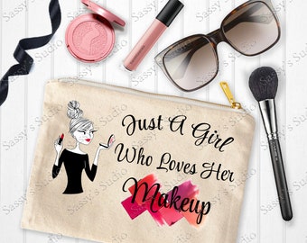 May include: A beige canvas makeup bag featuring a black and white illustration of a woman with a bun, holding lipstick and a compact mirror. The bag has the text "Just A Girl Who Loves Her Makeup" in black with red and pink brushstrokes.