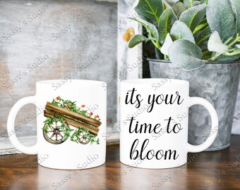 May include: Two white ceramic mugs. One mug features a watercolor illustration of a wooden wheelbarrow overflowing with green vines and red flowers. The other mug has the text "its your time to bloom" in black script.