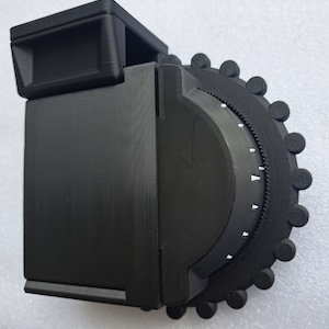 May include: A black 3D printed combination lock with a circular dial and a rectangular body. The dial has a series of numbers and a small arrow pointer.