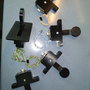 May include: Five black joystick controllers with round buttons and wires. The controllers are on a gray surface.