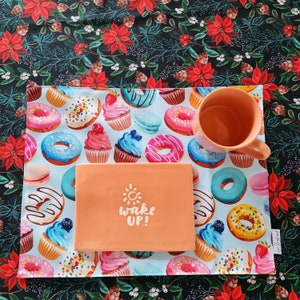 Könnte beinhalten: Eine Tischmatte mit hellblauem Hintergrund mit bunten Donuts und Cupcakes. Ein orangefarbener Becher und ein Teller mit dem Text "wake up!" befinden sich auf der Tischmatte. Der Hintergrund ist ein dunkles Blumenmuster.