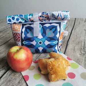 Könnte beinhalten: Eine weiße Lunch-Tasche mit einem geometrischen Muster in Blau, Orange und Grün. Die Tasche ist gefaltet und hat einen weißen Riemen. Ein roter Apfel und drei goldbraune Gebäckstücke liegen auf einer weißen Serviette mit bunten Punkten.