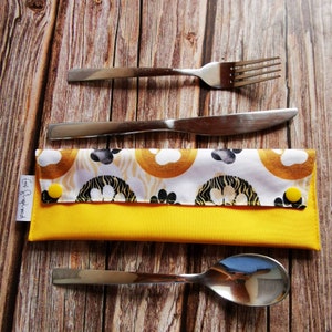 May include: A yellow fabric cutlery roll with a white and gold patterned fabric flap. The roll has two yellow snaps and the text "Eat & Repeat" printed on the side. A fork, knife, and spoon are placed on a wooden surface next to the roll.