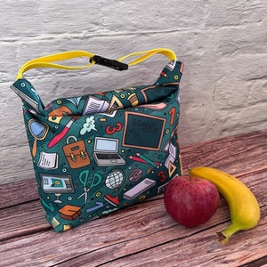 May include: A teal lunch bag with a yellow strap and black buckle, decorated with a school-themed pattern including a laptop, scissors, and a chalkboard with the word "SCHOOL". A red apple and a banana are placed beside the bag.