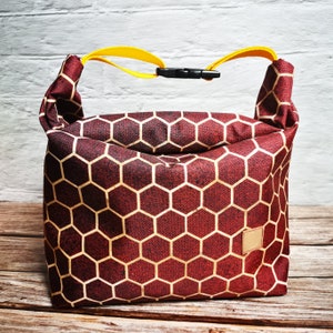 Könnte beinhalten: Eine rote und goldene Lunch-Tasche mit einem Wabenmuster und einem gelben Griff. Die Tasche hat einen schwarzen Reißverschluss.
