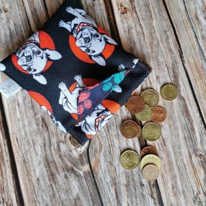 Könnte beinhalten: Ein schwarzes Stoff-Münzbeutel mit einem sich wiederholenden Muster aus weißen Bulldoggen-Illustrationen auf orangefarbenen Kreisen. Der Beutel ist offen und zeigt ein paar Münzen. Der Hintergrund ist eine Holzoberfläche.