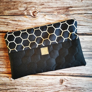 Könnte beinhalten: Schwarze Reißverschlusstasche mit einem goldenen Wabenmuster auf der oberen Klappe und einem gesteppten schwarzen Stoff auf der Unterseite. Die Tasche hat ein kleines goldenes Etikett mit einem Design.
