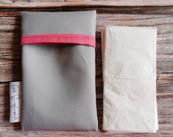 Può includere: Un astuccio in tessuto grigio con una fascia rosa e un'etichetta bianca con testo. Accanto, un fazzoletto bianco piegato. L'astuccio è rettangolare e sembra essere realizzato in un materiale resistente. Il fazzoletto ha una fascia strutturata.
