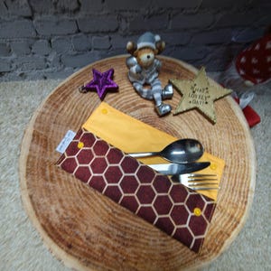 May include: A yellow and burgundy honeycomb pattern cutlery holder containing a spoon, knife, and fork. The holder is on a wooden slice with decorative stars and a figurine.
