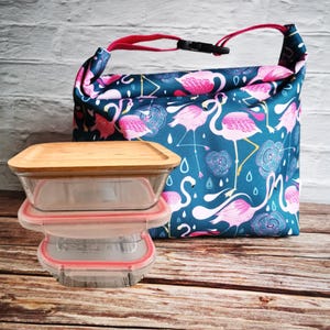 May include: A blue lunch bag with a flamingo print and pink accents. The bag is made of fabric and has a zipper closure. A set of three clear glass containers with bamboo lids are in front of the bag.