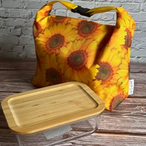 Op de afbeelding: Een gele lunchtas met een zonnebloemprint en een zwarte gesp. Voor de tas staat een glazen container met een bamboe deksel.