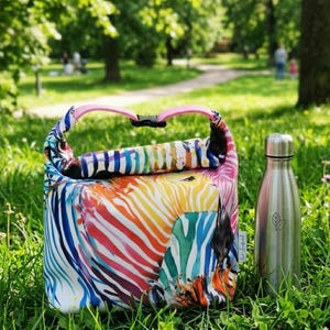 Op de afbeelding: Een kleurrijke geïsoleerde lunchtas met een levendig zebra-streepmotief in regenboogtinten, met een roze handvat en zwarte gesp. Een roestvrijstalen waterfles met een bladlogo staat ernaast.