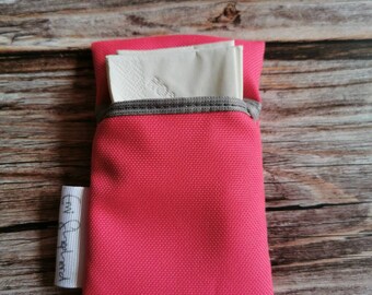 Può includere: Un porta fazzoletti in tessuto rosa acceso con una fascia elastica grigia, contenente fazzoletti bianchi. È attaccata una piccola etichetta bianca con testo nero. Il porta fazzoletti è su una superficie di legno.