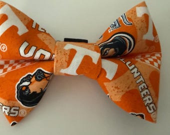May include: An orange bow tie featuring a repeating pattern of the white letter T, the word VOLUNTEERS, and a mascot graphic. The fabric has a speckled texture and includes small white checkered accents throughout the design of the accessory.