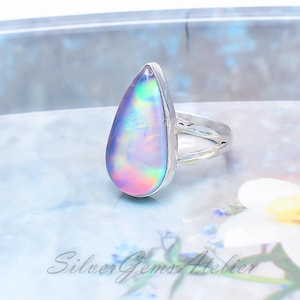 Puede incluir: Un anillo de plata con una gran piedra iridiscente en forma de lágrima con una superficie lisa y pulida que muestra colores cambiantes en rosa, azul y verde. La banda es de plata pulida y presenta un diseño dividido en los laterales.