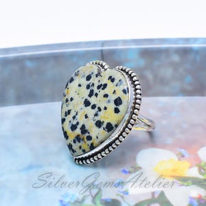 May include: A heart-shaped ring featuring a speckled dalmatian jasper stone with black spots on a cream and yellow background. The stone is set in a polished silver bezel with a beaded edge detail. The band is made of smooth, polished silver metal.