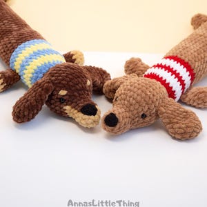 May include: Two crocheted dachshund plush toys made of soft brown yarn. One dog wears a blue and yellow striped sweater, while the other wears a red and white striped sweater. Both have black noses and eyes. The text AnnasLittleThing is visible.