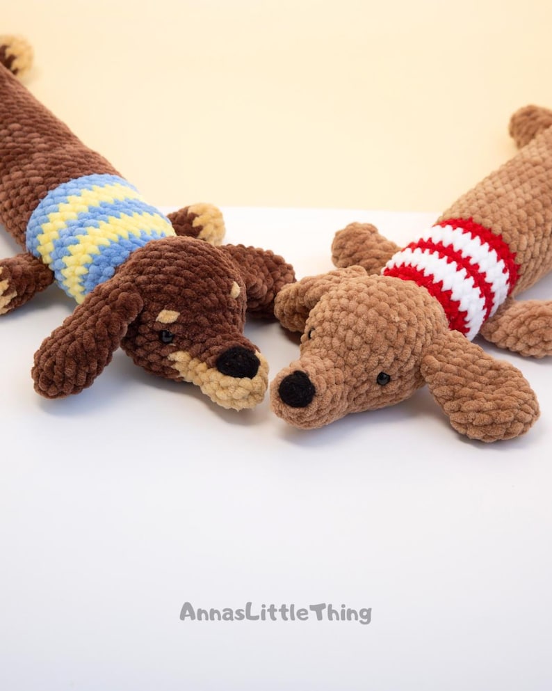 May include: Two crocheted dachshund plush toys made of soft brown yarn. One dog wears a blue and yellow striped sweater, while the other wears a red and white striped sweater. Both have black noses and eyes. The text AnnasLittleThing is visible.