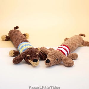 May include: Two crocheted dachshund plush toys made of soft brown yarn. One dog features a blue and yellow striped sweater, while the other wears a red and white striped sweater. Both have small black eyes and noses. The text AnnasLittleThing is visible.