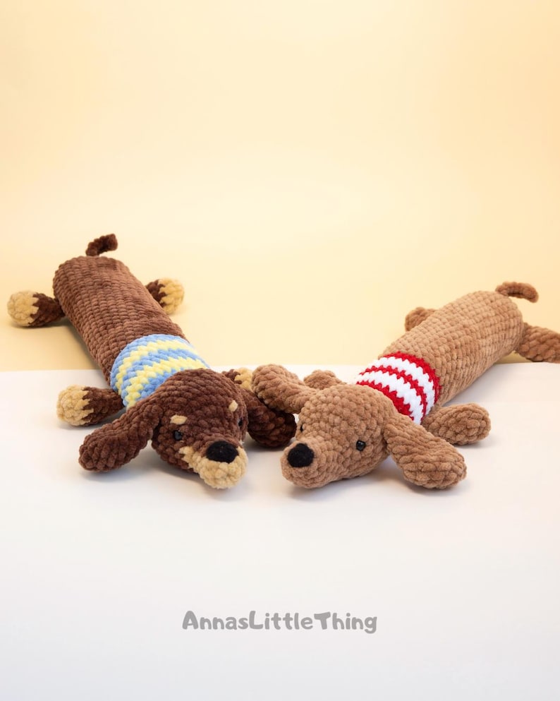 May include: Two crocheted dachshund plush toys made of soft brown yarn. One dog features a blue and yellow striped sweater, while the other wears a red and white striped sweater. Both have small black eyes and noses. The text AnnasLittleThing is visible.