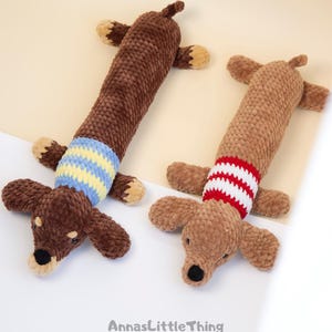 May include: Two plush dachshund toys made of textured brown yarn. The left toy is dark brown with a blue and yellow striped sweater. The right toy is light brown with a red and white striped sweater. The text AnnasLittleThing is at the bottom.
