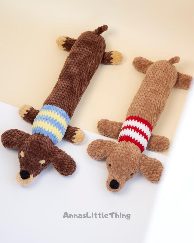 May include: Two plush dachshund toys made of textured brown yarn. The left toy is dark brown with a blue and yellow striped sweater. The right toy is light brown with a red and white striped sweater. The text AnnasLittleThing is at the bottom.