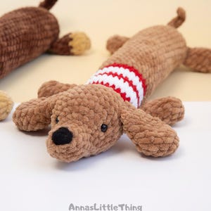 May include: Two crocheted plush dachshund toys made from soft brown yarn. The foreground dog features a red and white striped sweater pattern around its torso, black yarn eyes, and a black nose. The text AnnasLittleThing is printed at the bottom.